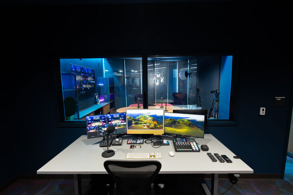 Podcast control room with a computer and audio control board with a window to the podcast studio.