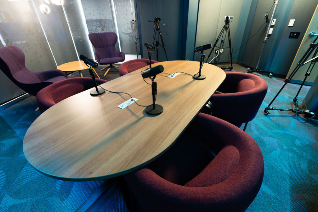 Podcast studio with colorful lighting, comfortable chairs around a table with microphones.