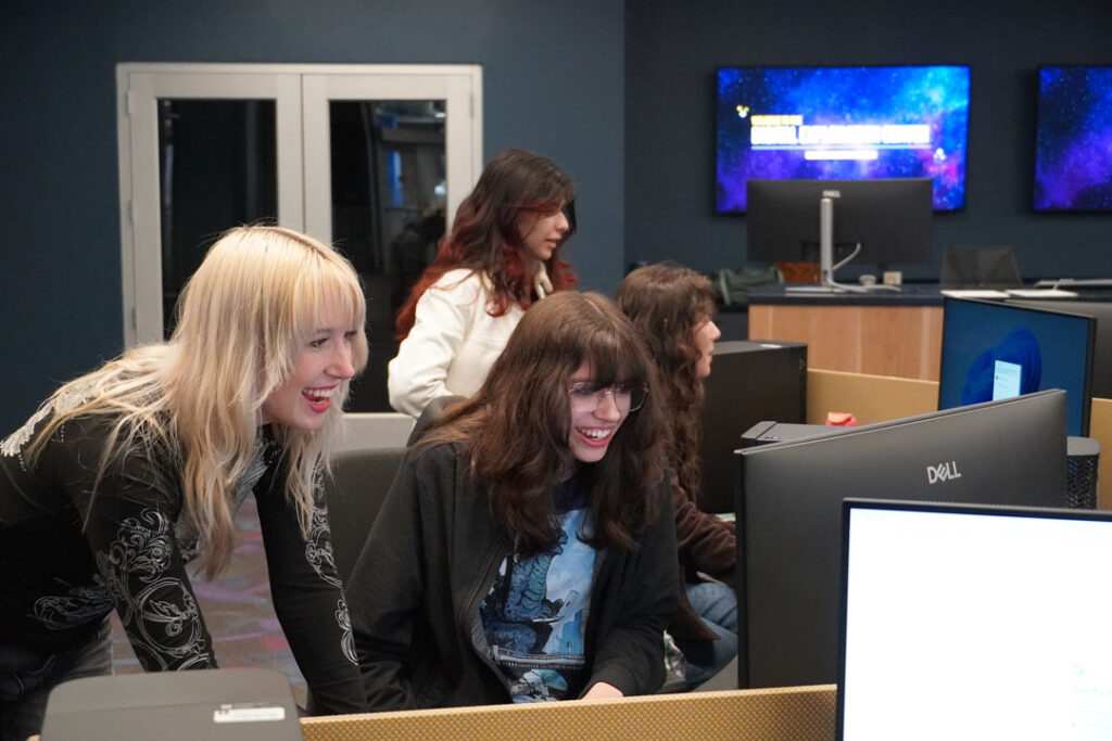 Female students sit around a computer workstation looking at a monitor.