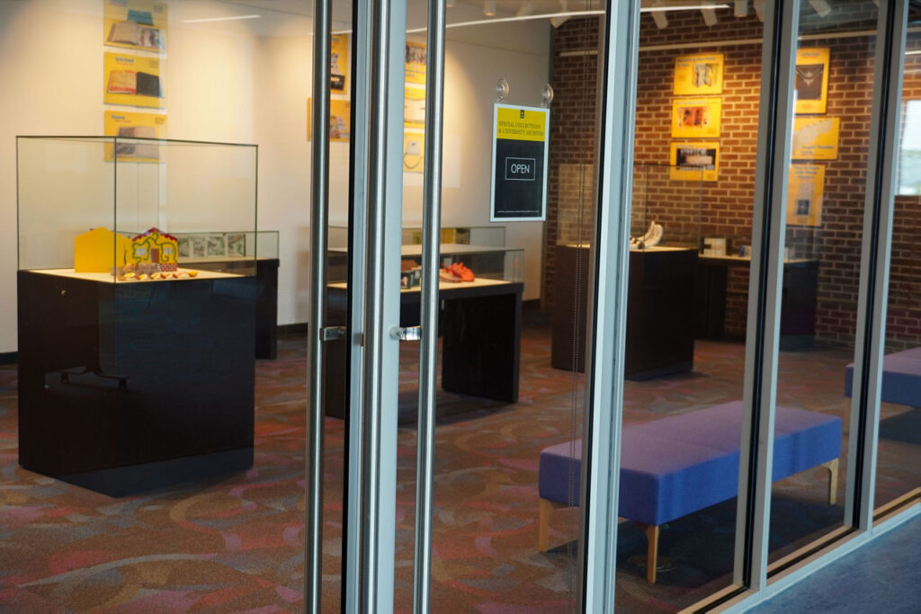 View into the exhibition through glass doors showing multiple display cases, a bench, and wall-mounted yellow exhibit panels; a sign on the door reads “OPEN.”