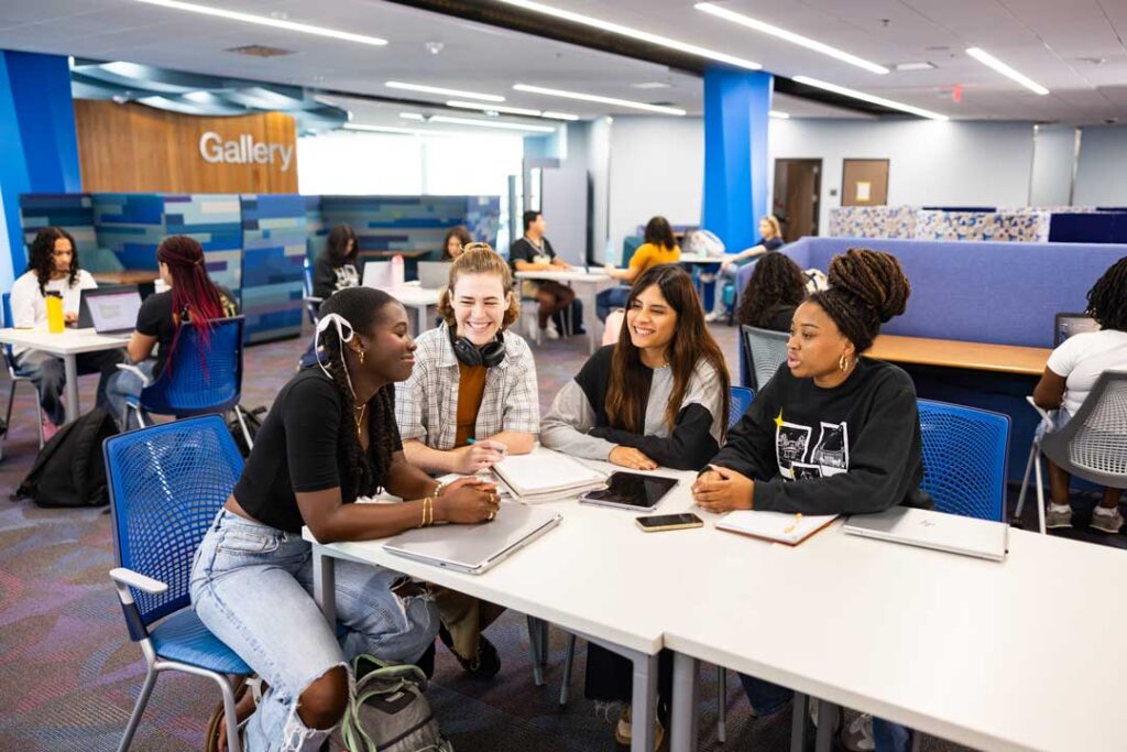 Students collaborate around tables in a bright library learning space, using laptops, tablets, and notebooks. Additional students study and work together throughout the room, with blue seating, collaborative work areas, and a "Gallery" sign visible in the background.