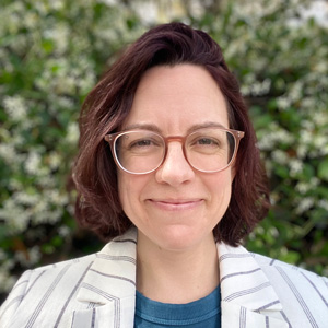 Head-and-shoulders portrait of Rae Mair with short dark hair and glasses, wearing a light striped blazer and blue top, standing outdoors in front of green foliage.
